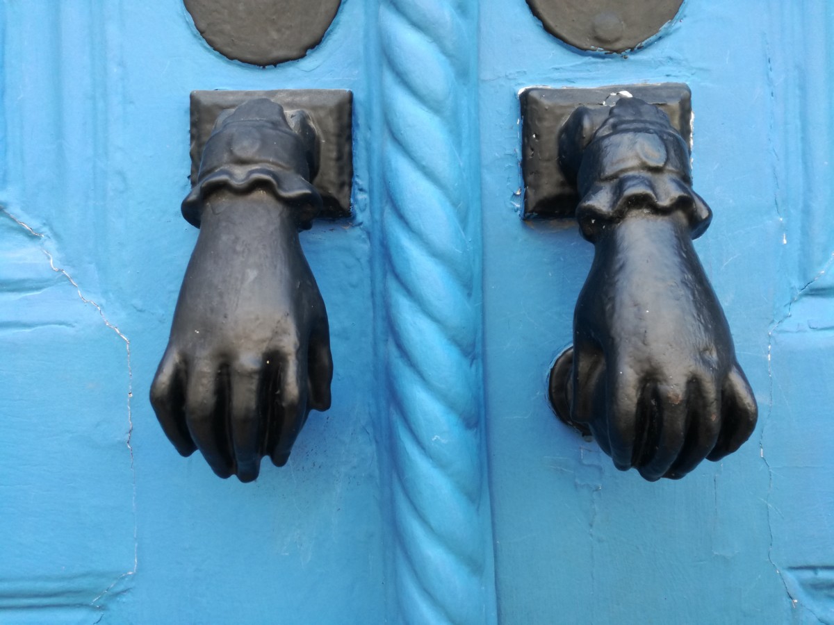 Een lichtblauwe deur met twee zwarte handjes met een ring aan de middelvinger en een kanten manchet.