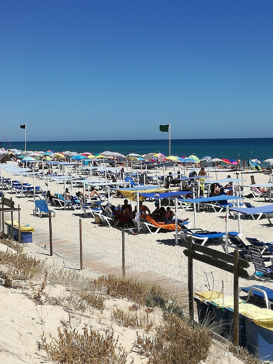 Strand van Barril in augustus 2018. Je ziet kleurige parasollen en daarachter een strakblauwe zee. De groene vlaggen geven aan dat zwemmen veilig is.