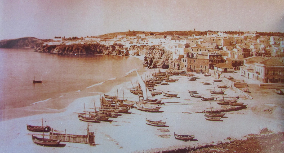 Een sepiakleurige foto van het strand van Albufeira in 1900. Er liggen veel boten op het strand, geen mensen.