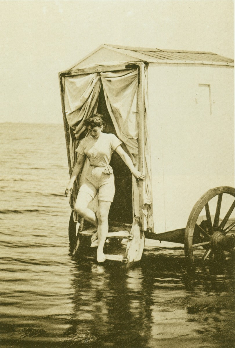 Een jonge vrouw in een zwempak dat haar schouders en bovenbenen bedekt, stapt uit een badkoetsje dat in ondiep water staat. Zwart-witfoto uit 1893.