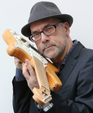 Foto van Peter Wessel die langs een gitaarhals in de camera kijkt. 
