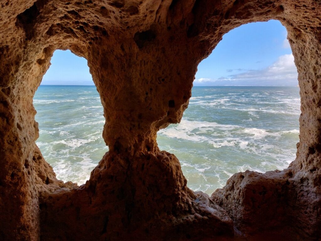 Twee ovale 'ramen' van een grot waardoor je een woeste blauwe schuimende oceaan ziet.