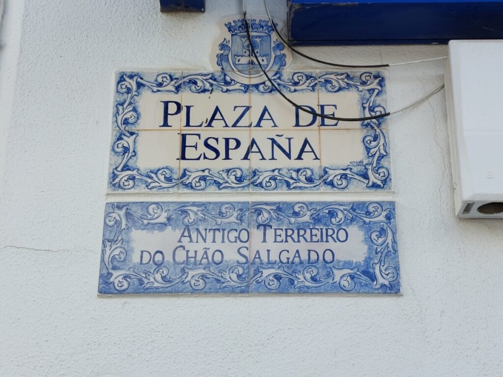 Straatnaambord met 'Plaza de España' en daaronder de in het Portugees: 'voormalig Terreiro do Chão Salgado'