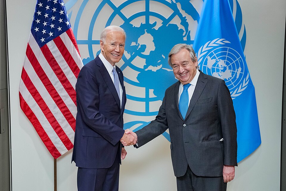 Guterres en Biden voor het logo van de Verenigde Naties en de vlaggen van de VN en de USA