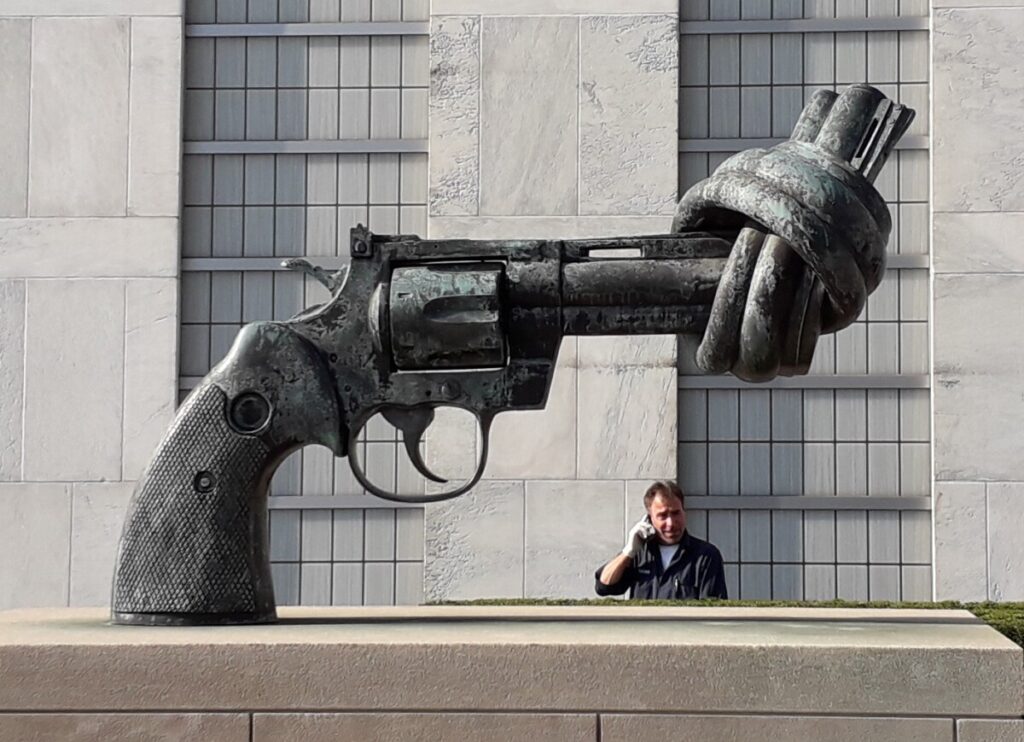 Beeldhouwwerk voor het gebouw van de VN, een revolver met een knoop in de loop. Op de achtergrond staat een man aan de telefoon te praten. Foto Cees Groenewegen